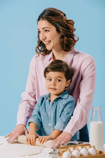 Feliz madre con adorable hijo rodando masa juntos aislado en azul - foto de stock