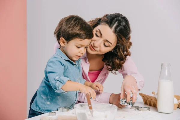 Feliz madre tiernamente mirando lindo pequeño hijo mientras cocina pastelería juntos sobre fondo bicolor - foto de stock