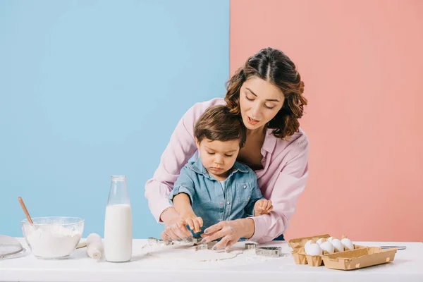 Pretty mother and little son cutting figures in dough together on bicolor background — Stock Photo