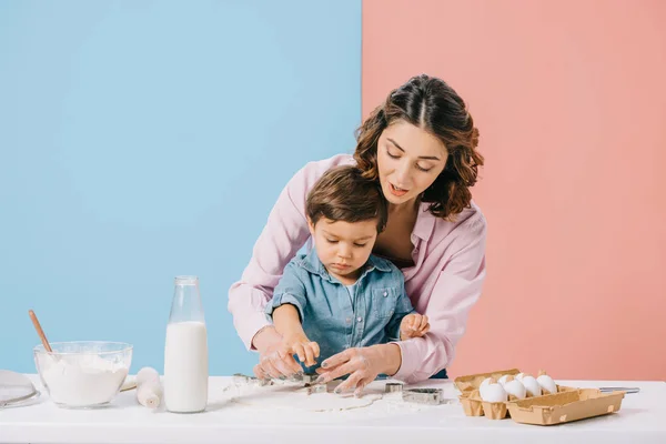 Hübsche Mutter und kleiner Sohn schneiden gemeinsam Figuren in Teig auf zweifarbigem Hintergrund — Stockfoto