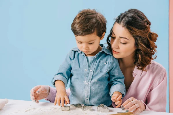 Mother and cute little son cutting figures in dough with dough molds together on bicolor background — Stock Photo