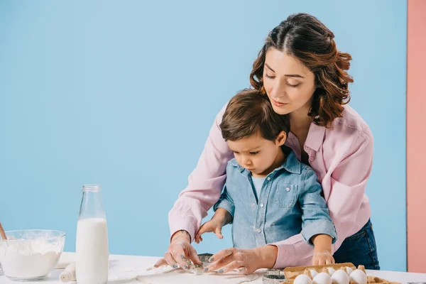 Madre y lindo hijo pequeño pastelería de cocina juntos en la mesa de la cocina blanca sobre fondo bicolor - foto de stock