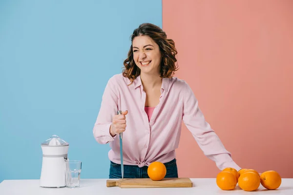 Mulher atraente segurando faca enquanto estava na mesa da cozinha com laranjas frescas no fundo bicolor — Fotografia de Stock