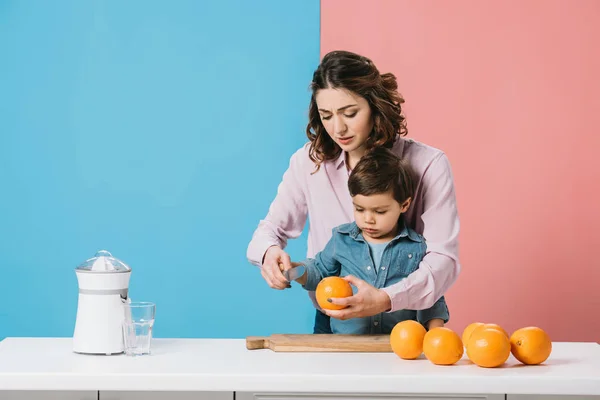 Lindo niño mirando naranja a la mano de las madres mientras está de pie junto a la mesa de la cocina con naranjas sobre fondo bicolor - foto de stock