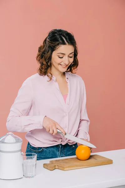 Lächelnd schöne Frau schneidet Orange auf Holzschneidebrett isoliert auf rosa — Stockfoto