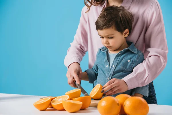 Милый маленький мальчик режет апельсины вместе с матерью изолированы на синий — стоковое фото