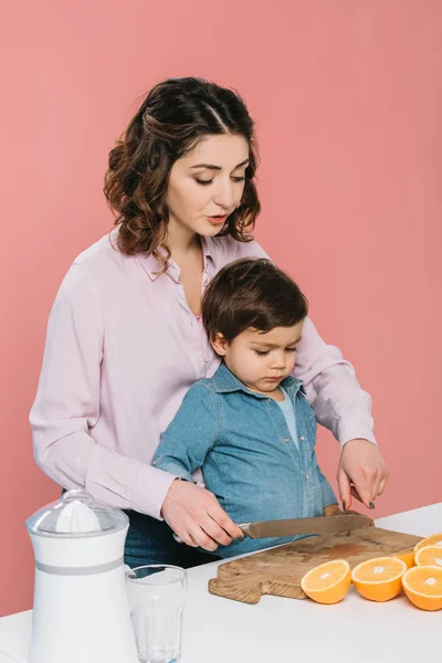 Madre mostrando naranjas cortadas a pequeño hijo mientras sostiene cuchillo aislado en rosa - foto de stock