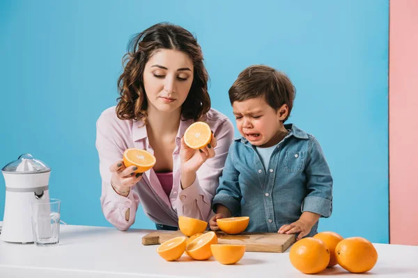 Mother holding cut oranges and little son crying while standing at kitchen table on bicolor background — Stock Photo