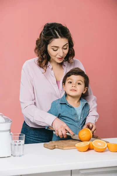 Glückliche Mutter mit niedlichem kleinen Sohn, der zusammen frische Orangen isoliert auf rosa schneidet — Stockfoto