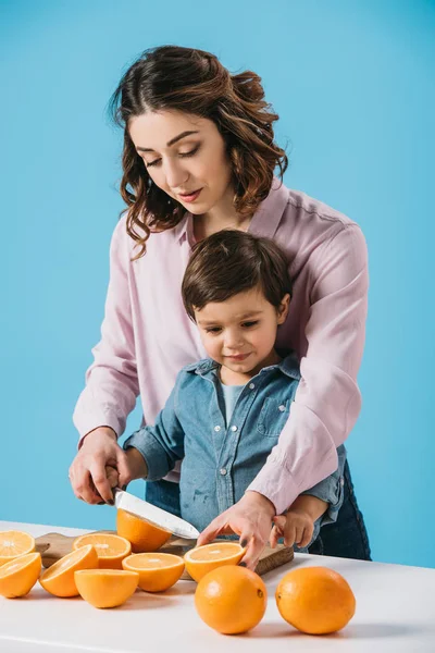 Jolie mère avec mignon petit fils oranges de coupe sur planche à découper en bois ensemble isolé sur bleu — Photo de stock