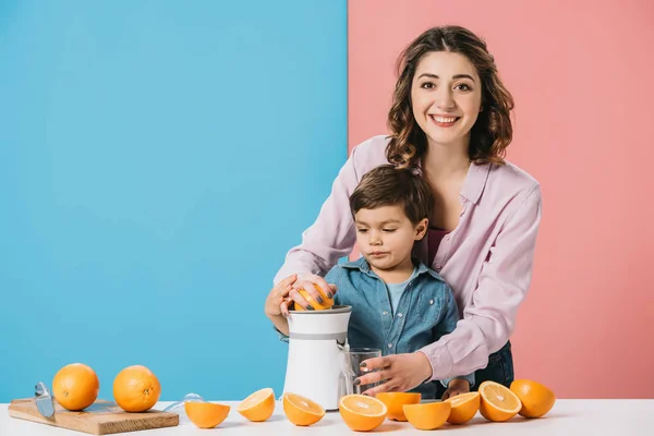 Heureux mère serrant jus d'orange frais avec mignon petit fils sur fond bicolore — Photo de stock