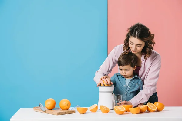 Мать с маленьким сыном сжимает апельсиновый сок на белом кухонном столе на двухцветном фоне — стоковое фото