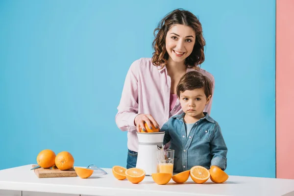 Усміхнена мати стискає сік разом з милим маленьким сином і дивиться на камеру на біколорному фоні — стокове фото