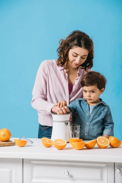 Lindo niño ayudando a la madre en exprimir jugo de naranja fresco aislado en azul - foto de stock