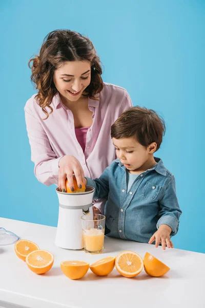 Улыбающаяся мать с милым маленьким сыном, сжимающим свежий апельсиновый сок на соковыжималке, изолированной на голубом — стоковое фото
