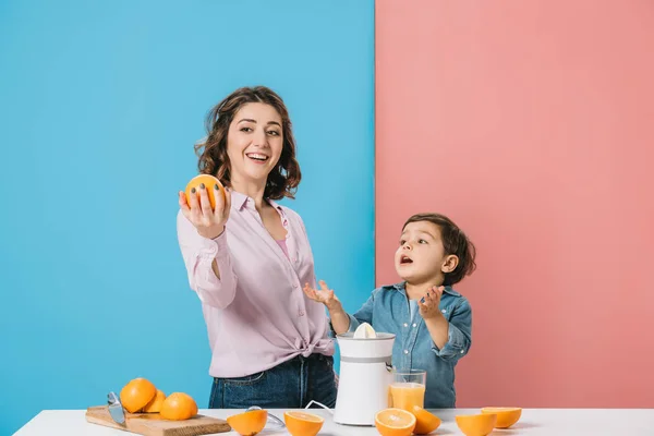 Happy mother showing whole fresh orange to cute little son stretching hands on bicolor background — Stock Photo