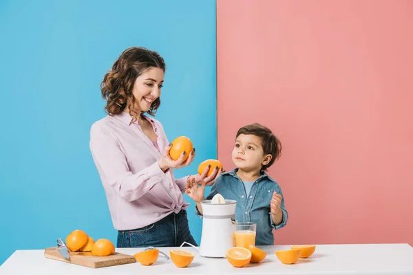 Glückliche Mutter zeigt ihrem niedlichen kleinen Sohn ganze Orangen auf zweifarbigem Hintergrund — Stockfoto
