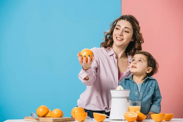 Smiling mother showing whole orange to cute little son on bicolor background — Stock Photo