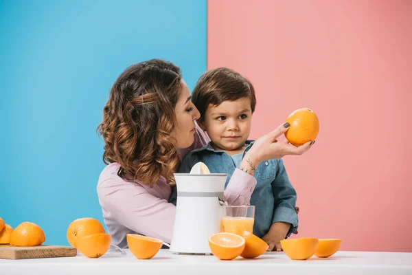 Mãe mostrando laranja fresca inteira para o filho pequeno bonito no fundo bicolor — Fotografia de Stock