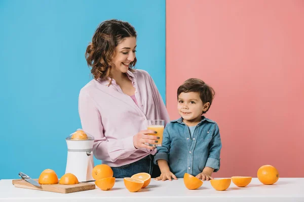 Sonriente madre dando completo vaso de jugo de naranja fresco a lindo hijo pequeño sobre fondo bicolor - foto de stock