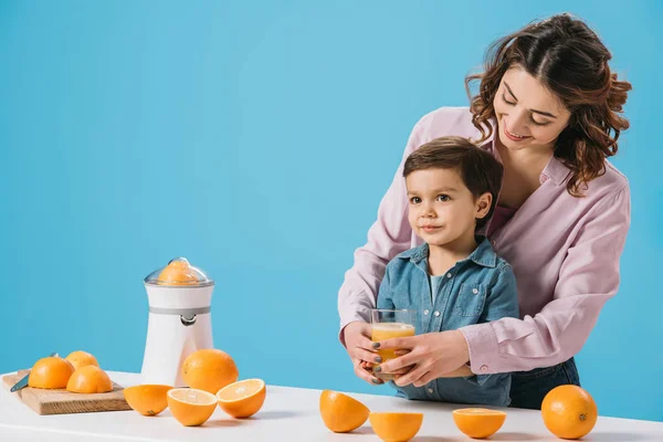 Sonriente madre dando completo vaso de jugo de naranja fresca a lindo pequeño hijo aislado en azul - foto de stock