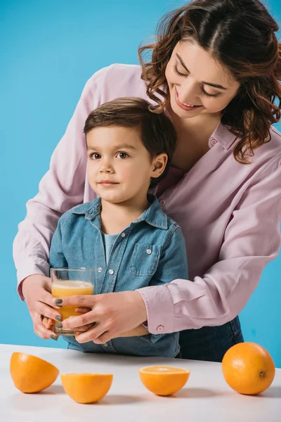Feliz madre dando completo vaso de jugo de naranja fresca a adorable pequeño hijo aislado en azul - foto de stock