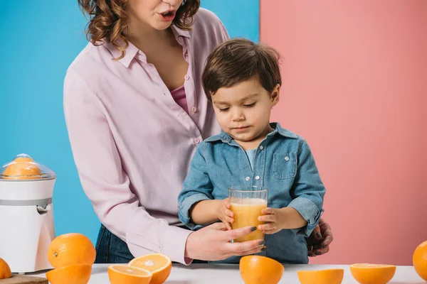 Carino bambino prendendo bicchiere pieno di succo d'arancia fresco da madri mano su sfondo bicolore — Foto stock