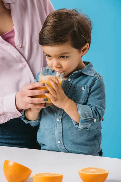 Cute holding glass with mothers hand and drinking fresh orange juice isolated on blue — Stock Photo