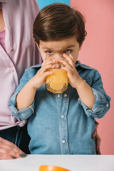 Niedlicher kleiner Junge trinkt frischen Orangensaft, während er mit Mutter am Küchentisch auf zweifarbigem Hintergrund steht — Stockfoto