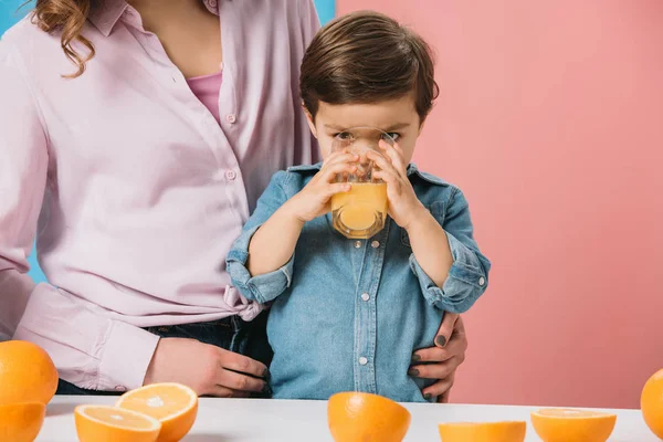 Adorável menino desfrutando de suco de laranja fresco enquanto de pé com a mãe por mesa de cozinha no fundo bicolor — Fotografia de Stock