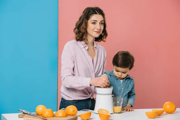 Glückliche Mutter mit niedlichem kleinen Sohn, der frischen Orangensaft auf Saftpresse zusammen auf zweifarbigem Hintergrund presst — Stockfoto