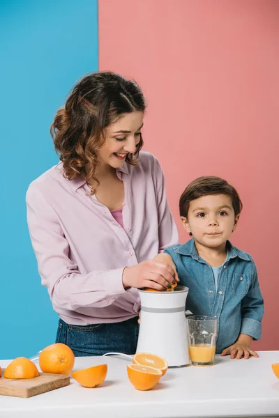 Усміхнена мати і чарівний маленький син стискає свіжий апельсиновий сік разом на біколорному фоні — стокове фото