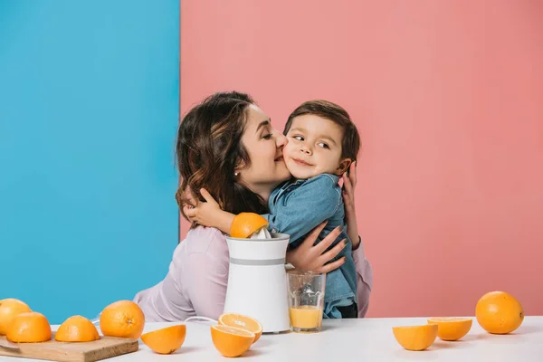 Happy mother hugging adorable smiling little son by kitchen table with fresh oranges and juicer on bicolor background — Stock Photo