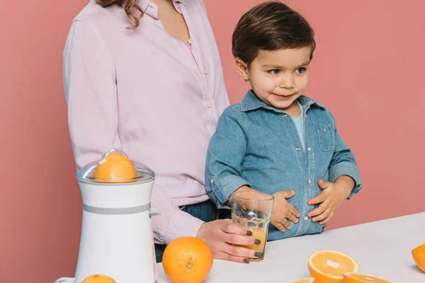 Cute little boy holding hands on tummy while standing at kitchen table with mother holding glass with fresh orange juice isolated on pink — Stock Photo