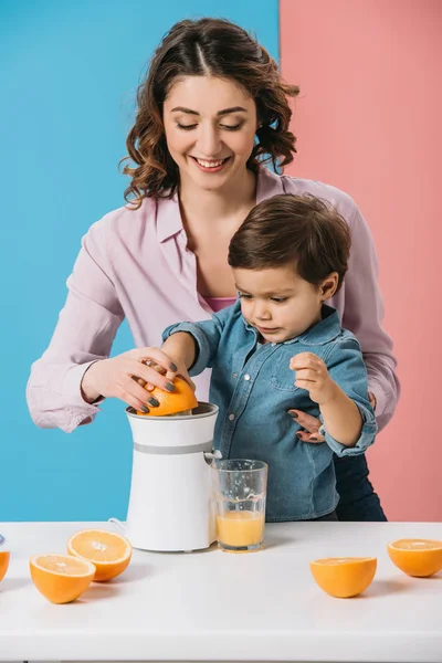 Счастливая мать с очаровательным сыном, сжимающим свежий апельсиновый сок вместе на двухцветном фоне — стоковое фото