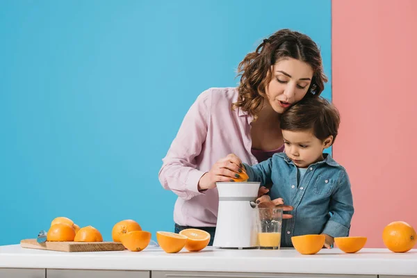 Милый маленький мальчик с матерью сжимая свежий апельсиновый сок на соковыжималке на двухцветном фоне — стоковое фото