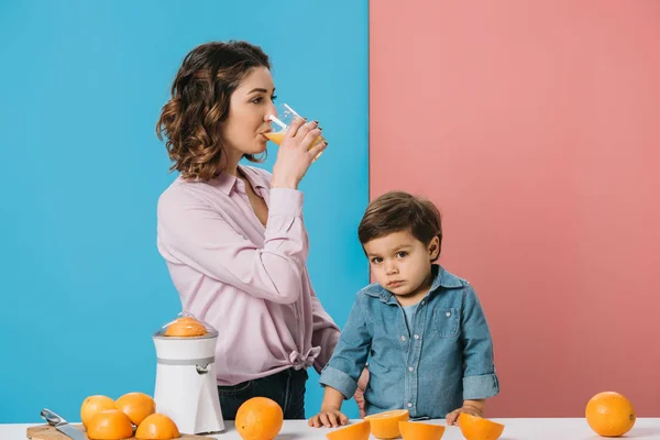 Mother drinking fresh orange juice while standing with cute little son by table with oranges on bicolor background — Stock Photo