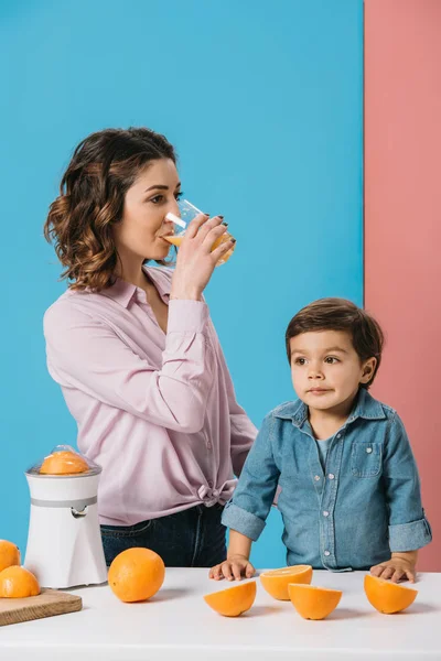 Madre godendo di succo d'arancia fresco mentre in piedi con adorabile piccolo figlio a tavola con arance su sfondo bicolore — Foto stock