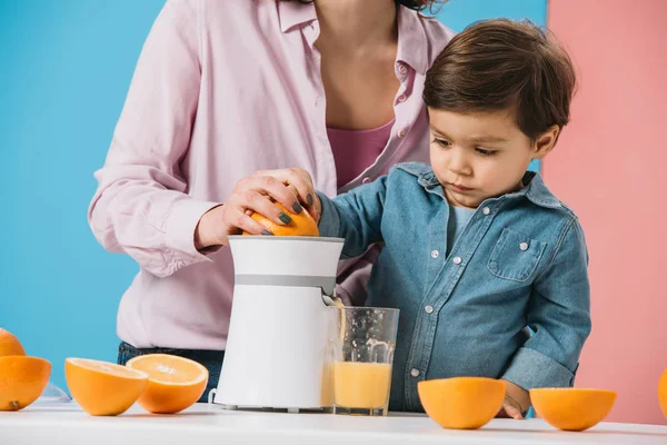 Adorable niño pequeño exprimiendo jugo de naranja fresco en exprimidor junto con la madre en fondo bicolor - foto de stock