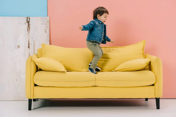 Mignon petit garçon en veste bleue et jeans verts sautant sur canapé jaune — Photo de stock