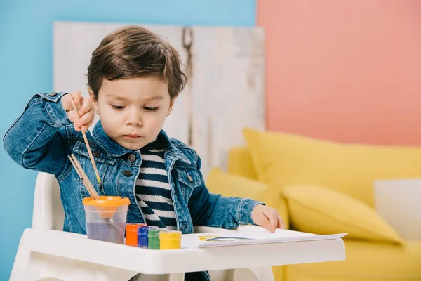 Netter kleiner Junge wählt Malpinsel, während er auf dem Hochstuhl mit Aquarellfarben auf dem Tisch sitzt — Stockfoto