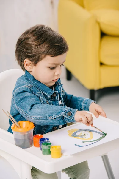 Adorable little boy painting with watercolor paints while sitting on highchair — Stock Photo