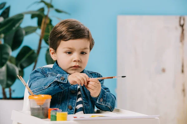 Lindo niño sosteniendo pincel de pintura mientras está sentado en la trona con pinturas de acuarela en la mesa - foto de stock