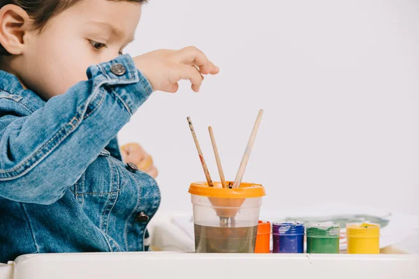 Cute little boy choosing painting brush while sitting on highchair with watercolor paints on table isolated on white — Stock Photo