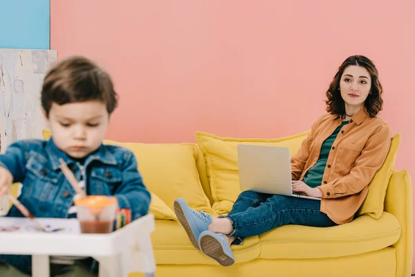 Mère assis sur un canapé jaune et utilisant un ordinateur portable tandis que mignon petit fils peinture à l'aquarelle — Photo de stock