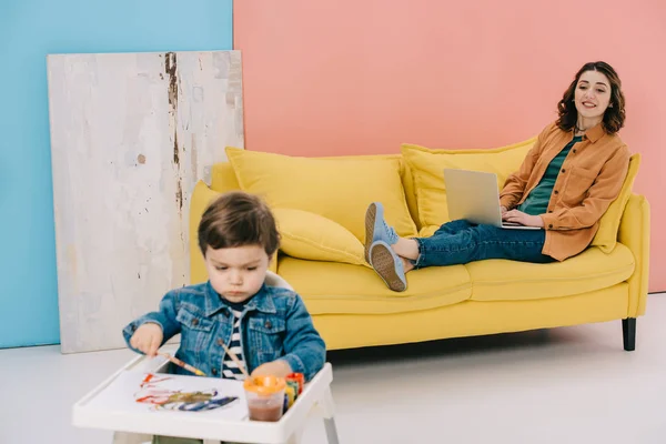Mãe sorridente sentado no sofá amarelo e usando laptop enquanto adorável pequena pintura filho com tintas aquarela — Fotografia de Stock