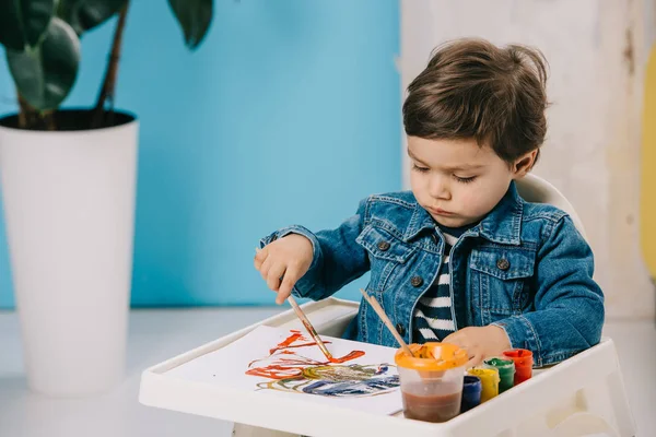 Bonito menino pintura com tintas aquarela enquanto sentado em cadeira alta — Fotografia de Stock