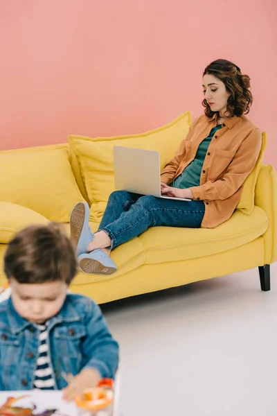 Mère assise sur un canapé jaune et utilisant un ordinateur portable tandis que le petit fils peint avec des peintures à l'aquarelle — Photo de stock