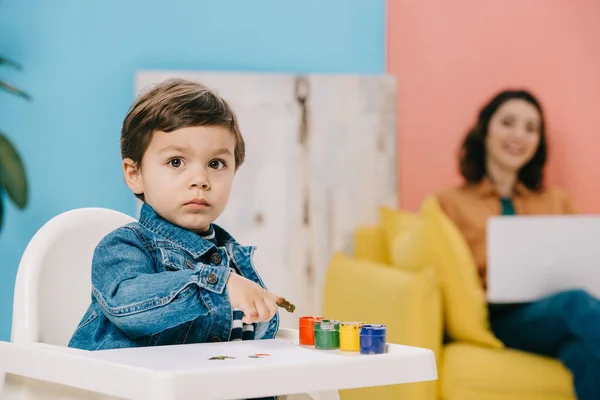 Cute little boy painting with watercolor paints with finger while mother sitting on yellow sofa and using laptop — Stock Photo