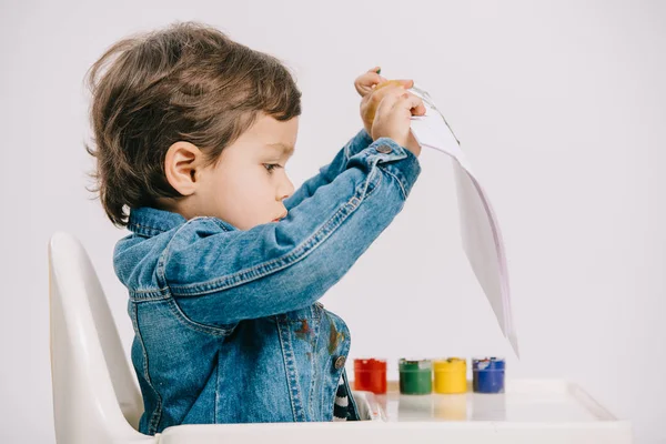 Cute little boy holding paper while sitting on highchair with watercolor paints on table isolated on white — Stock Photo